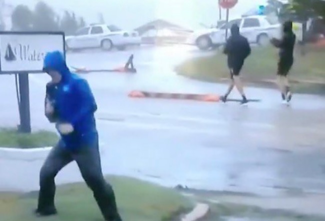 Image: Hilarious fake news: Weather Channel reporter fakes hurricane-force winds while two guys casually stroll down the street behind him, wearing shorts